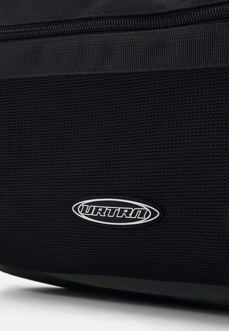 Bolsa de tela negra con un panel de malla texturizada, que presenta una insignia de logo plateada. Incluye una cremallera negra de contraste por encima de la malla.