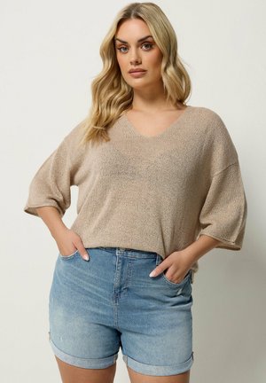 Blondynka ubrana w beżowy, luźny sweter z dzianiny z rękawami do łokcia oraz jasnoniebieskie podwinięte dżinsowe szorty, stoi z rękami w kieszeniach.