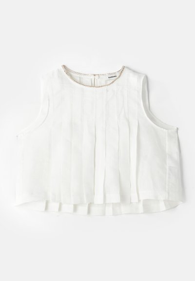 Fehér ujj nélküli blúz redőkkel, kerek nyakkivágással, amelyet fémes díszítés és gombos zárás díszít a hátán.