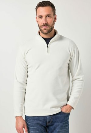 Man met baard, gekleed in een wit fleece pullover met kwart-ritssluiting en blauwe spijkerbroek, staand met één hand in de zak tegen een effen achtergrond.