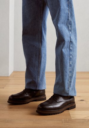 Scarpe stringate in pelle marrone scuro indossate con jeans blu a taglio dritto su pavimento in legno chiaro contro parete semplice.