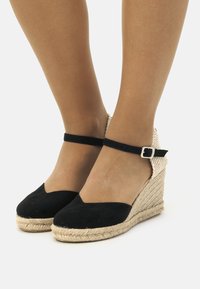 Zapatos de cuña de ante negro con una suela de cuerda de yute. Cuentan con una correa en el tobillo con una hebilla plateada y un diseño de punta redonda.