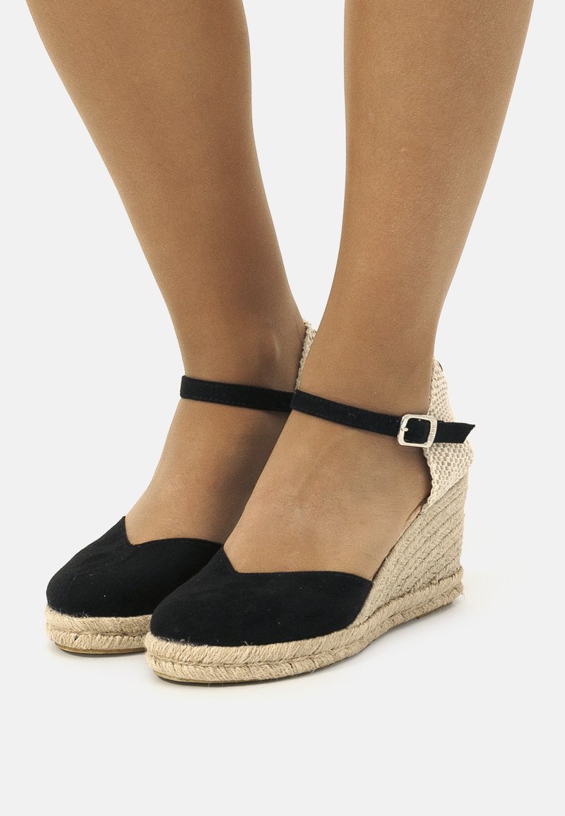 Zapatos de cuña de ante negro con una suela de cuerda de yute. Cuentan con una correa en el tobillo con una hebilla plateada y un diseño de punta redonda.