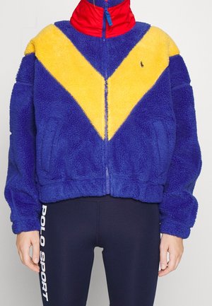 Fleecejacke mit blauem Körper, gelbem Chevron auf der Brust, rotem Kragen, durchgehendem Reißverschluss, Seitentaschen, kombiniert mit marineblauen Leggings mit der Aufschrift "Polo Sport".