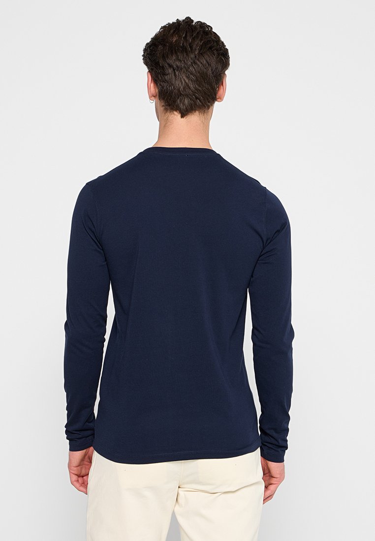 Mann mit kurzen braunen Haaren, der ein langärmliges, marineblaues Hemd und hellfarbene Hosen trägt, steht mit dem Gesicht von der Kamera abgewandt vor einem schlichten Hintergrund.