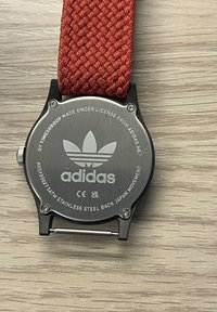 Sarkans auduma siksna pulkstenis ar nerūsējošā tērauda aizmuguri, kas piedāvā Adidas zīmolu un gravētas detaļas par licenci un kustību.