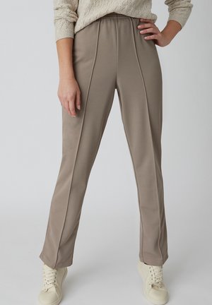 Donna che indossa pantaloni taupe a gamba dritta con cuciture centrali, un maglione color crema e sneakers avorio con lacci, in piedi con mano sul fianco.