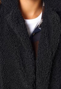 Svart, texturerad fleecejacka med ett mjukt, fluffigt utseende och öppet framdesign, lager på en vit skjorta.