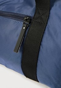 Μπλε σακίδιο από νάιλον με μαύρο λουρί και κλείσιμο με φερμουάρ. Υφασμάτινη υφή με κομψό, μινιμαλιστικό σχεδιασμό. Ορατές μαύρες μεταλλικές λεπτομέρειες.