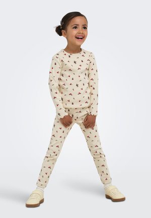 Ung pige står med benene fra hinanden iført cremefarvet top og bukser med små røde blomsterprint og beige sko.