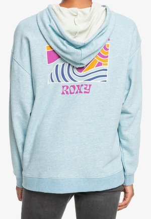 Personne portant un sweat à capuche bleu clair avec un motif abstrait coloré ondulé et le texte « ROXY » au dos, associé à un pantalon foncé.
