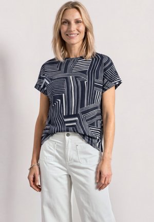 Femme blonde souriante portant un top à manches courtes imprimé géométrique noir et blanc et un pantalon blanc taille haute sur un fond uni.