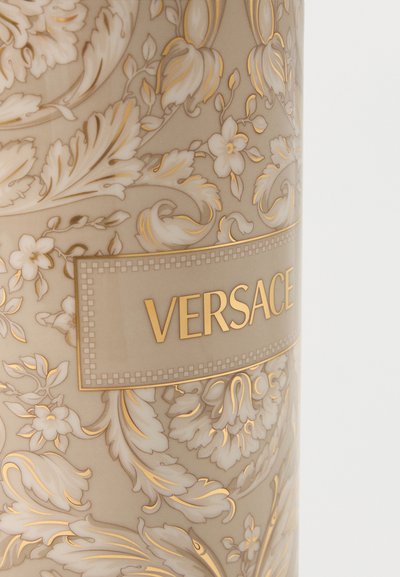 Surface à motif floral beige et or avec le mot « VERSACE » en lettres capitales dorées centré sur une étiquette rectangulaire.