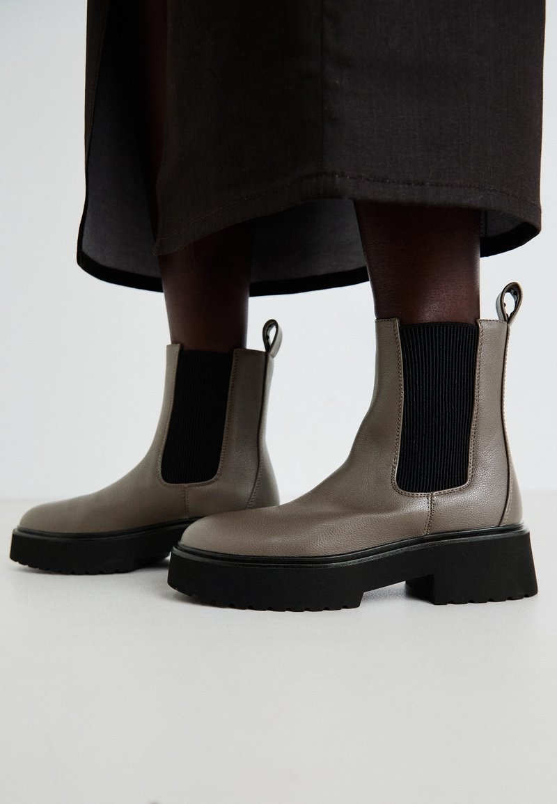 Botines de cuero gris con paneles elásticos en los lados, suela negra gruesa y lengüeta trasera para facilitar el calzado. Acabado texturizado en el cuero.