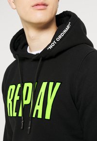 Sudadera negra con letras audaces en neón verde que dicen "REPLAY". La capucha tiene impresa en el borde una frase contrastante en blanco que dice "NO ORDINARIO".