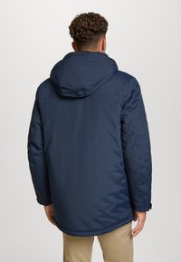 Giacca imbottita blu navy con cappuccio, caratterizzata da una superficie liscia, orlo sul retro leggermente più lungo e polsini regolabili. Design semplice senza motivi.