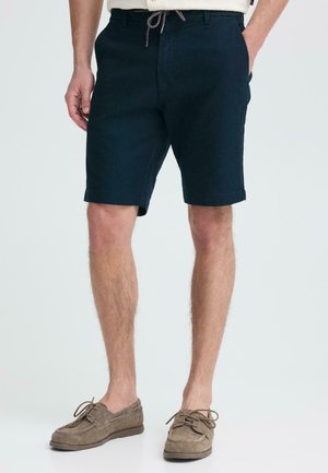 Man draagt marineblauwe shorts met trekkoord en zijzakken, bruin suède veterschoenen en staat voor een effen achtergrond.