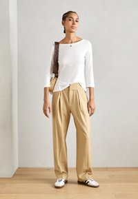 Top blanco de manga larga, pantalones beige con pliegues y zapatillas blancas con rayas negras. Bolsa marrón al hombro con tirante estampado.