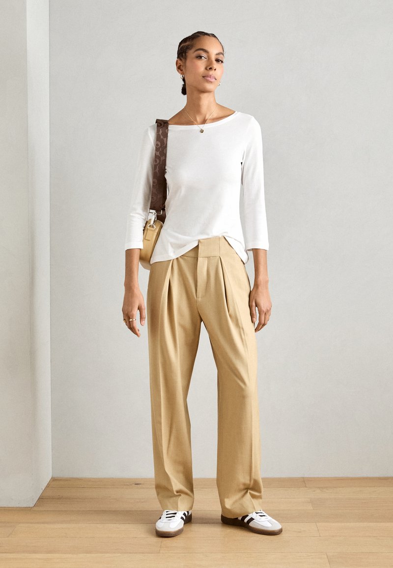 Top blanco de manga larga, pantalones beige con pliegues y zapatillas blancas con rayas negras. Bolsa marrón al hombro con tirante estampado.