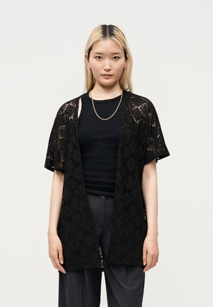 Junge Person mit glatten blonden Haaren trägt eine schwarze Spitzen-Open-Cardigan über einem schwarzen Top und dunkelgrauen Hosen vor einem einfarbigen hellen Hintergrund.