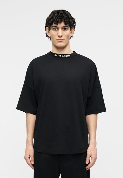 Bărbat tânăr cu păr creț, purtând un tricou negru larg cu textul "Palm Angels" pe guler, stând în picioare pe un fundal simplu și deschis la culoare.