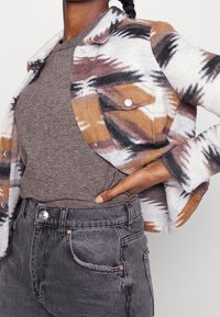 Button-up Karojacke mit braunen, weißen und schwarzen Mustern; weicher Stoff; kombiniert mit einem grauen Pullover und dunklen Jeans.