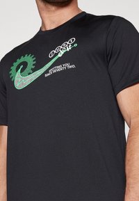 Černé tričko s krátkým rukávem, na kterém je zelený grafický design s textem "just do it" a "SPOTTING YOU SINCE SEVENTY TWO." v bílé barvě.