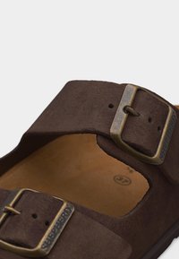 Καφέ σαουέν σανδάλι με δύο ρυθμιζόμενες ορειχάλκινες αγκράφες και ταμπά εσωτερική σόλα, μέγεθος 37, εμφανιζόμενο σε κοντινή λήψη σε λευκό φόντο.