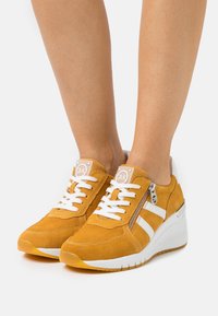 Żółte zamszowe sneakersy z białymi paskami i sznurówkami, z bocznym zamkiem błyskawicznym oraz podeszwą na koturnie dla zwiększenia wysokości. Teksturowana cholewka zapewniająca przyczepność.