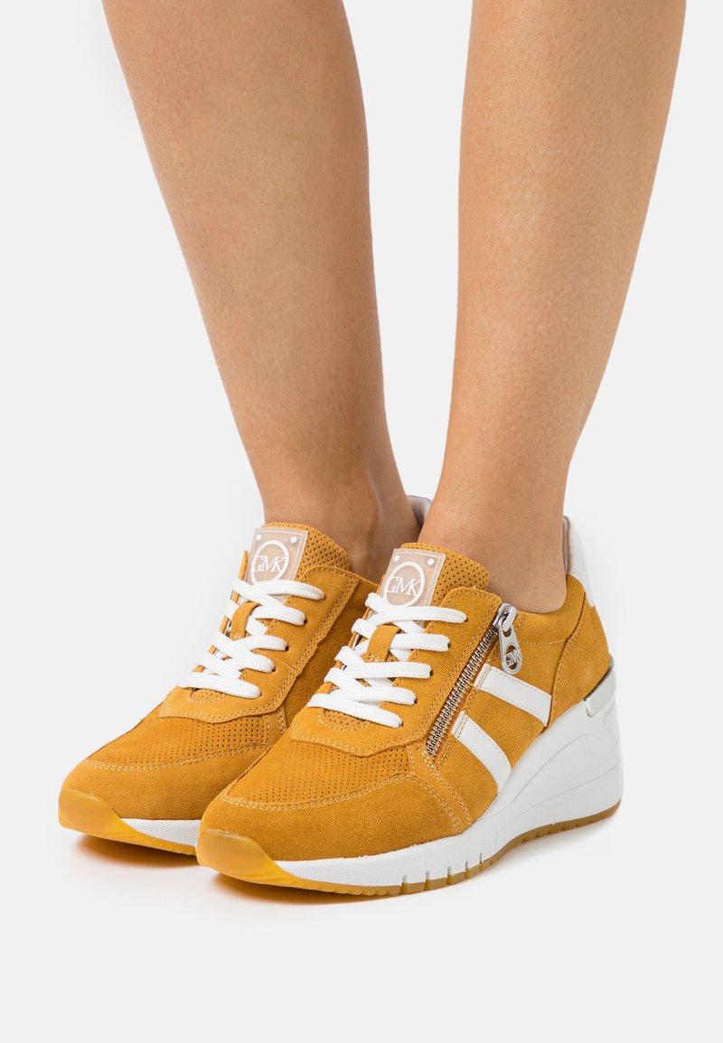 Żółte zamszowe sneakersy z białymi paskami i sznurówkami, z bocznym zamkiem błyskawicznym oraz podeszwą na koturnie dla zwiększenia wysokości. Teksturowana cholewka zapewniająca przyczepność.