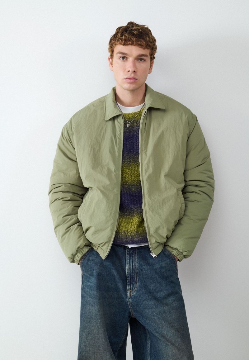 Giovane uomo con capelli ricci indossa una giacca imbottita verde chiaro, un maglione a righe e jeans blu larghi, in piedi contro un muro bianco.
