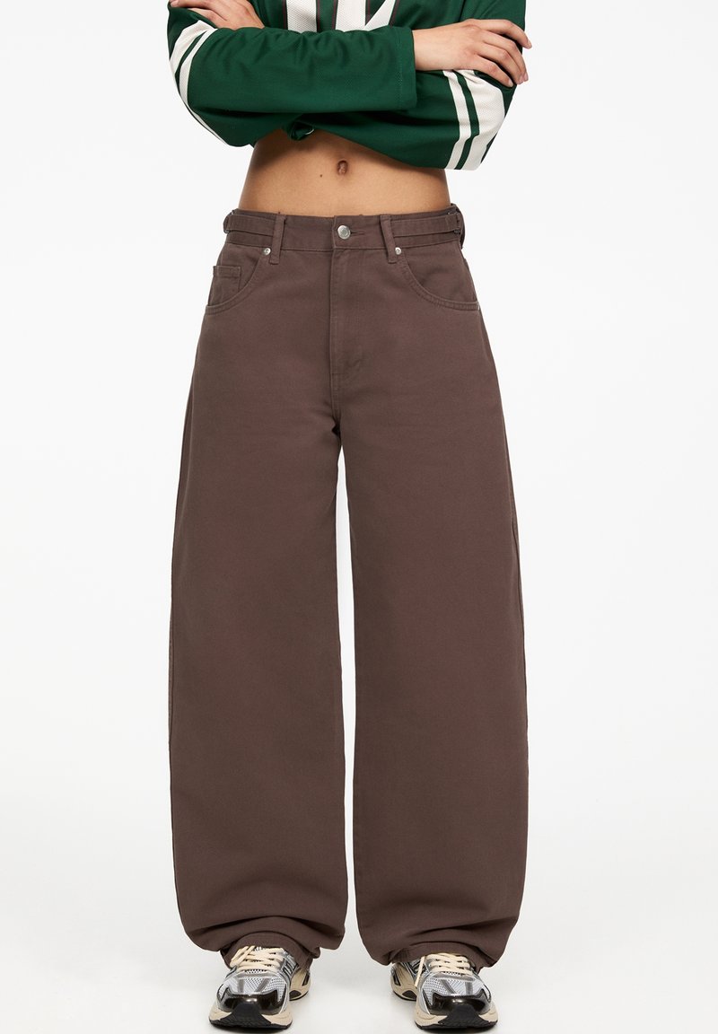 Personne portant un pantalon large marron ample, un haut court vert à manches longues et des baskets blanches, debout les bras croisés.