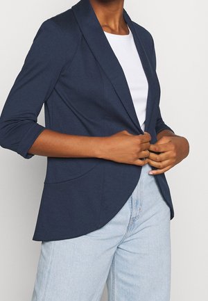 Person wearing a navy blazer over a white shirt and light blue jeans, adjusting the blazer with both hands.