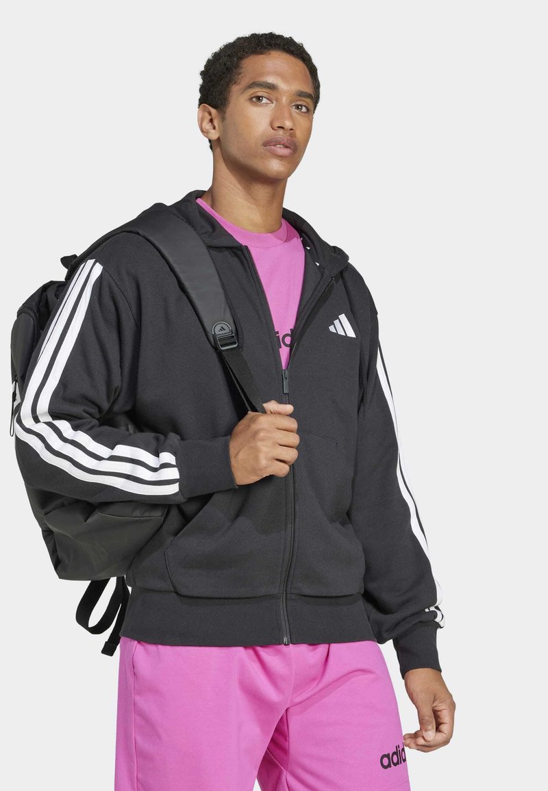 Black zip-up hoodie with white stripes on sleeves, worn over a pink shirt and paired with pink pants. Includes a black backpack.