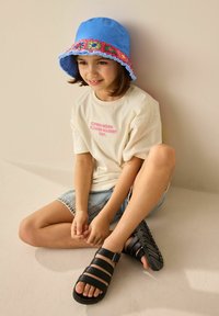 Hellbeige T-Shirt mit pinkem Text, blauer Bucket Hat mit buntem Stickmuster, Jeansshorts und schwarzen Sandalen mit Riemen.