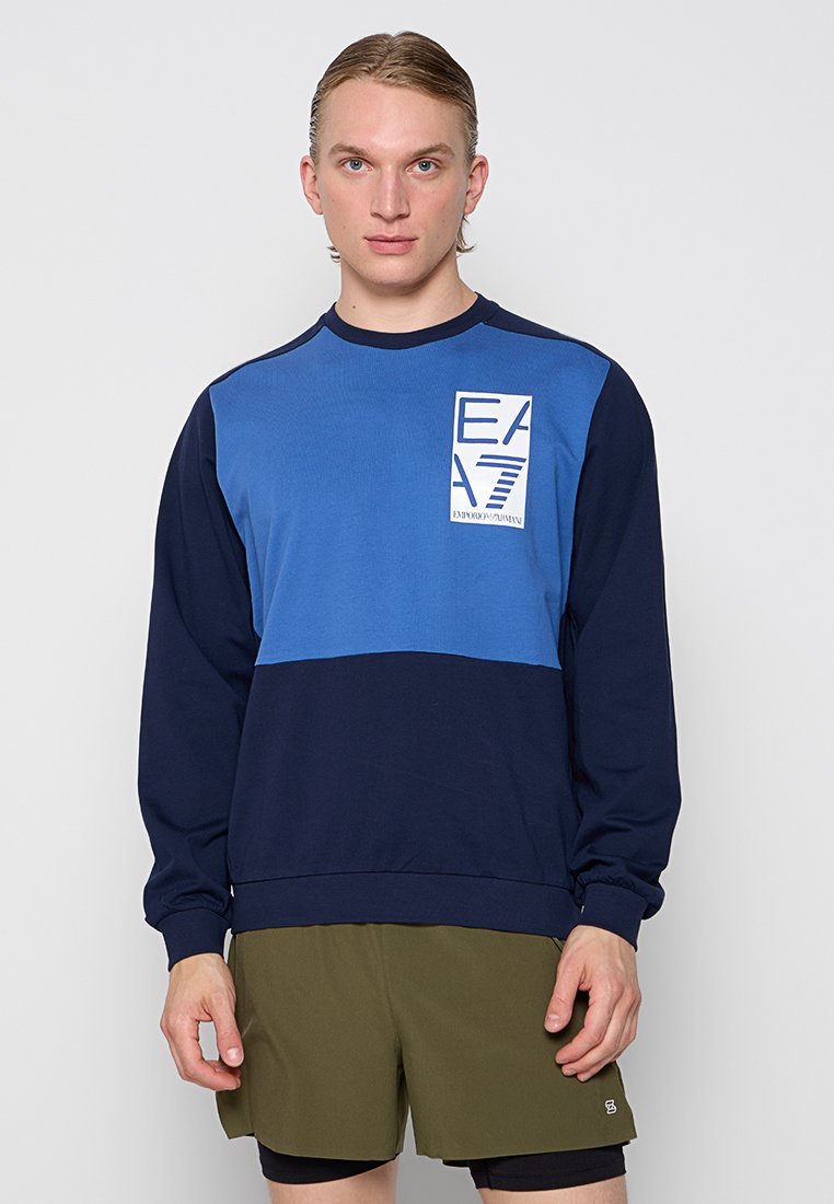 Joven con sudadera en bloques de color azul marino y azul con el logo de EA7, y pantalones cortos en verde oliva, de pie contra un fondo claro y liso.