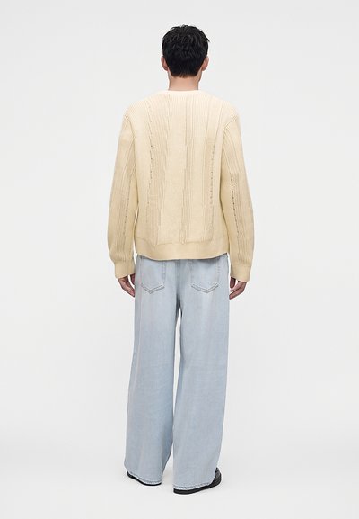 Homme debout dos à la caméra portant un pull en maille crème, un jean large bleu clair et des chaussures noires sur un fond uni.