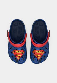 Blue rubber clogs with a red strap, featuring a Superman logo. The surface has comic-style patterns with various superhero-themed graphics.