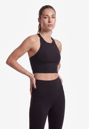 Frau steht mit den Händen in der Hüfte, trägt ein schwarzes ärmelloses Crop-Top und schwarze Leggings mit hoher Taille vor einem schlichten weißen Hintergrund.