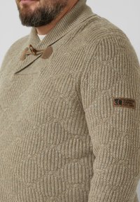 Beige gebreide trui met een gestructureerd patroon, voorzien van een sjaalkraag, houten klepsluiting en een zwart merkembleem op de linker mouw.