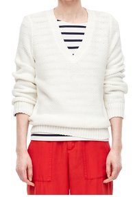 Pull blanc en maille avec un col en V et des manches retroussées, présentant un motif texturé. Porté sur un haut rayé et associé à un pantalon rouge.