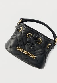 Μαύρο τσαντάκι χειρός από δερμάτινο καπιτονέ υλικό με χρυσό λογότυπο, στρογγυλή λαβή και χρυσά οριγκάλι. Διαθέτει υφή με παιχνιδιάρικη σχεδιαστική λεπτομέρεια.