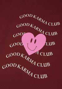 Bordeaux stof met herhalende witte tekst "GOOD KARMA CLUB" en een grote roze hart met een lachend gezicht in het midden.