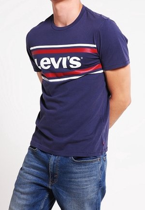Mann trägt ein marineblaues Levi's T-Shirt mit roten und weißen Streifen, kombiniert mit blauen Jeans, steht mit den Händen hinter dem Rücken.