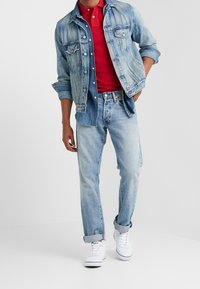 Jasnoniebieska kurtka dżinsowa nałożona na czerwoną koszulkę polo, w zestawieniu z jasnoniebieskimi dżinsami i białymi trampkami. Kurtka ma detale w postaci guzików i podwinięte rękawy.