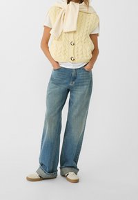 Żółta dzianinowa kamizelka z teksturowanym wzorem i dwoma guzikami, noszona na białej koszuli. Stylizację dopełniają jasnoniebieskie dżinsy o szerokich nogawkach i beżowe trampki.