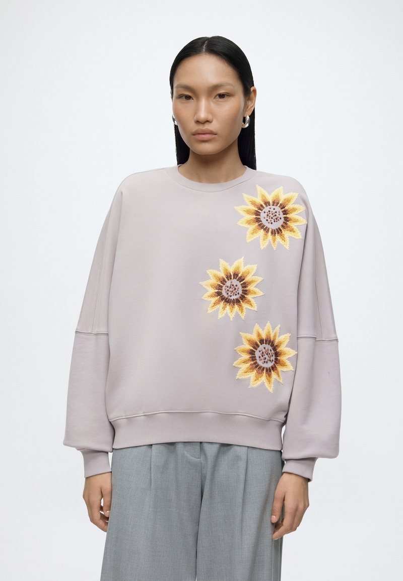 Femme portant un sweat-shirt violet clair avec trois grands tournesols brodés sur le devant et un pantalon gris à jambes larges.
