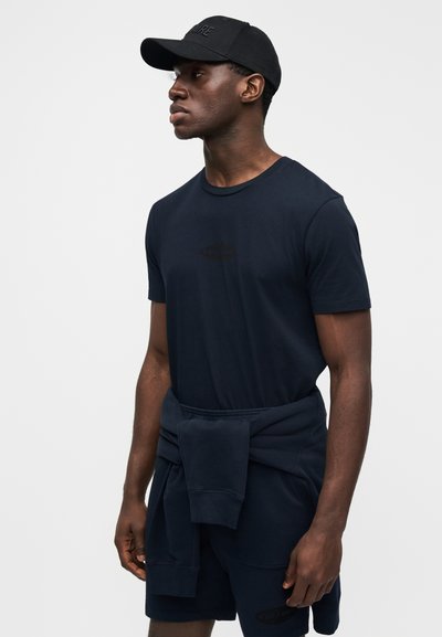 Μπλε σκούρο T-shirt με κοντό μανίκι και διακριτικό λογότυπο, σε συνδυασμό με ταιριαστά σορτς και μαύρο καπέλο. Το ύφασμα φαίνεται απαλό και άνετο.