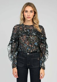 Blouse noire transparente à motifs floraux avec des volants superposés, des manches longues et un col froncé ; associée à un jean en denim foncé.