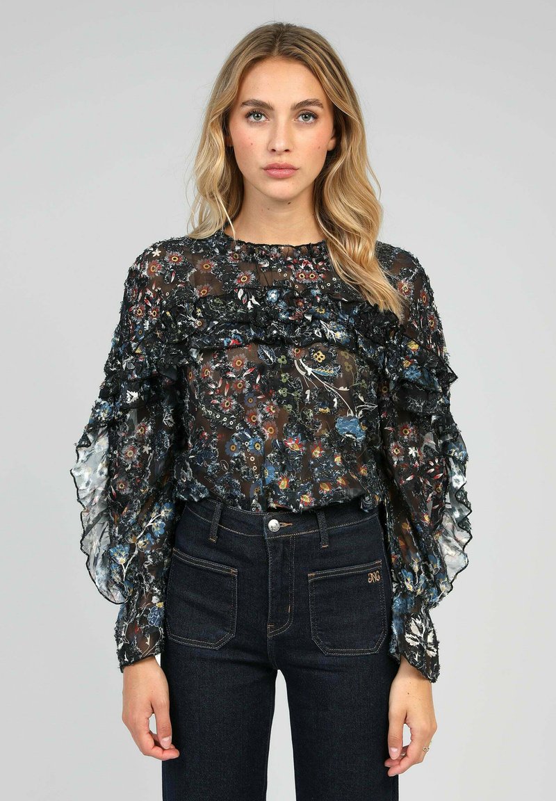 Blouse noire transparente à motifs floraux avec des volants superposés, des manches longues et un col froncé ; associée à un jean en denim foncé.